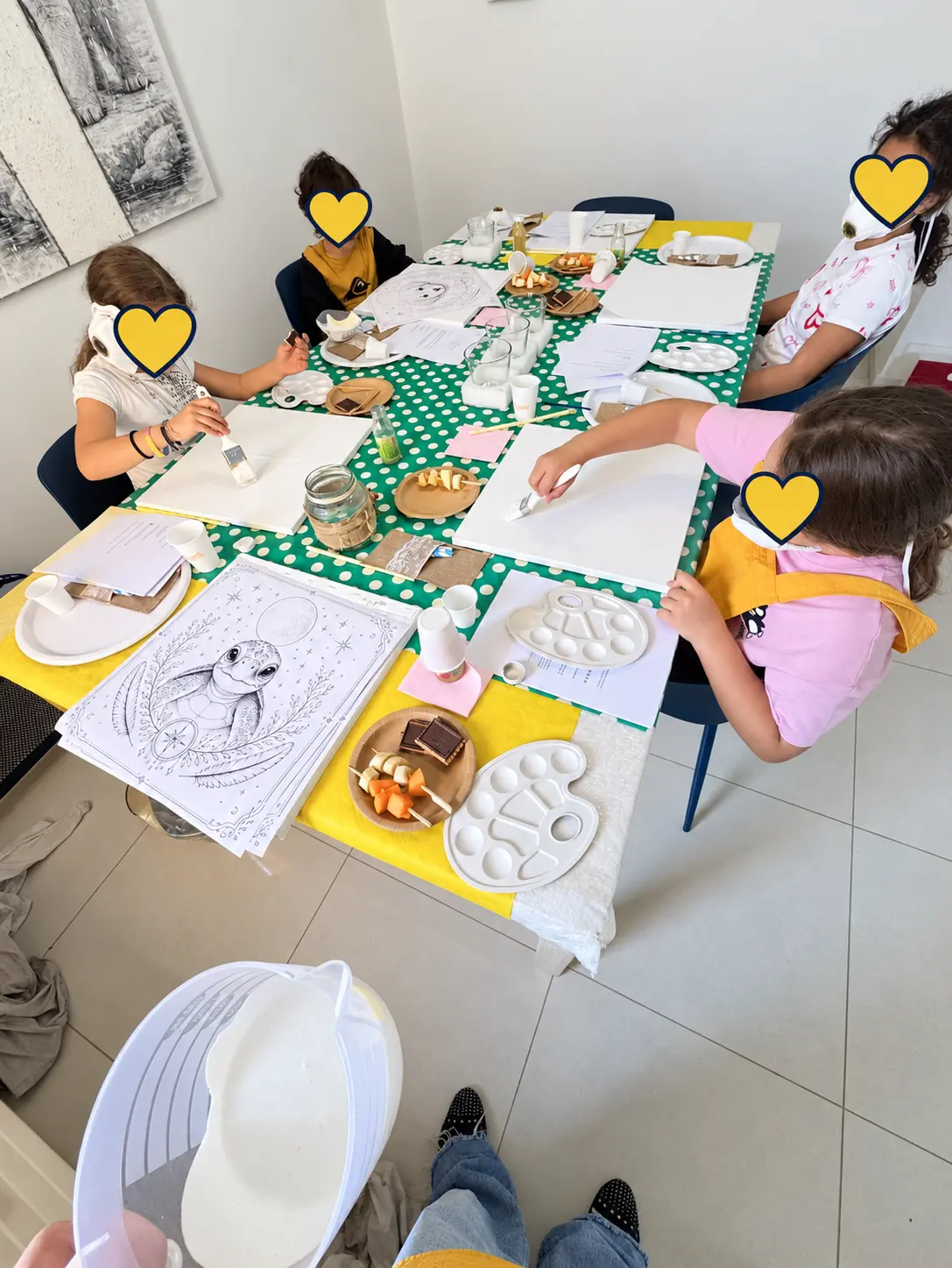 Quattro bambini dipingono insieme durante una Merenda con DAMA; i volti sono protetti da cuori gialli